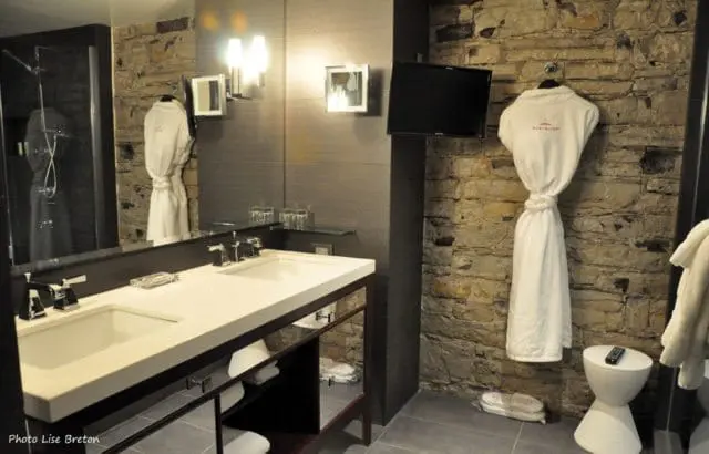 Modern bathroom with a double vanity, stone accent wall, and a hanging white bathrobe. A flat-screen TV is mounted on the stone wall.