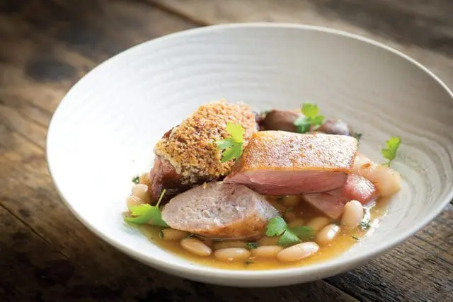 Cassoulet with duck confit, sausage, and white beans in a white bowl on a wooden table.