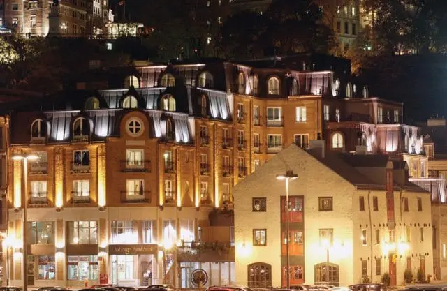 Multi-story brick buildings with mansard roofs illuminated at night, featuring arched windows and balconies. A lighter stone building with red doors is in the foreground.