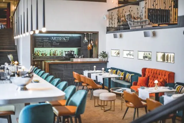Modern restaurant interior with a long white bar, teal and brown seating, and a mezzanine level with decorative railings. A chalkboard menu lists 'Artefact' and 'Huitres & Cocktails'.