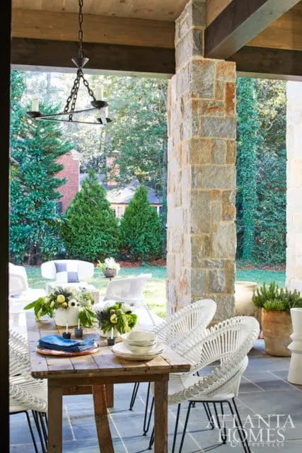 Covered porch with a rustic wooden dining table set with white plates and blue napkins, surrounded by white wicker chairs. A stone pillar and chandelier are visible.