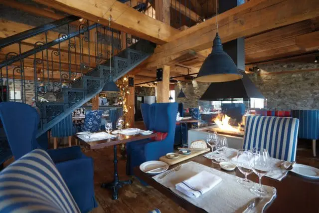 Restaurant dining room with blue upholstered chairs, dark wood tables, a large stone fireplace with a fire, and a wrought iron staircase.