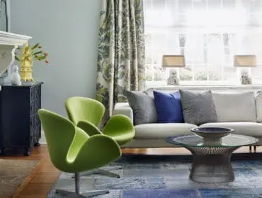 Living room with a white sectional sofa, lime green egg chairs, a dark wood dresser, and a fireplace. Patterned curtains hang by a large window.