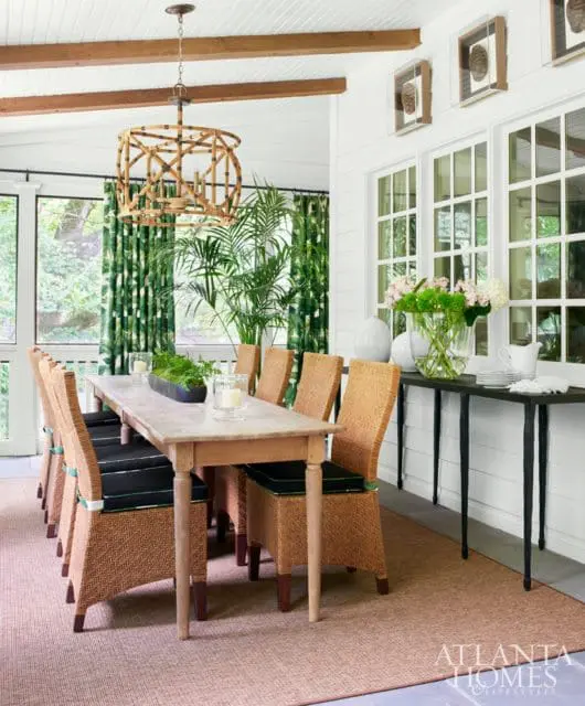 Dining room with a long wooden table surrounded by wicker chairs with black cushions, a bamboo chandelier, and green floral curtains.
