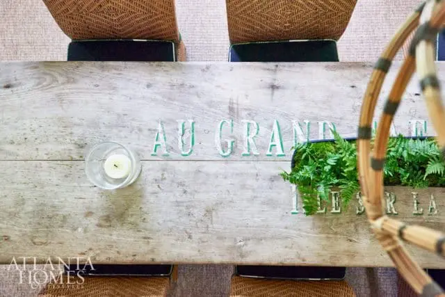 Rustic wooden dining table with green painted text reading 'AU GRAND', a glass votive with a white candle, and a planter filled with ferns.