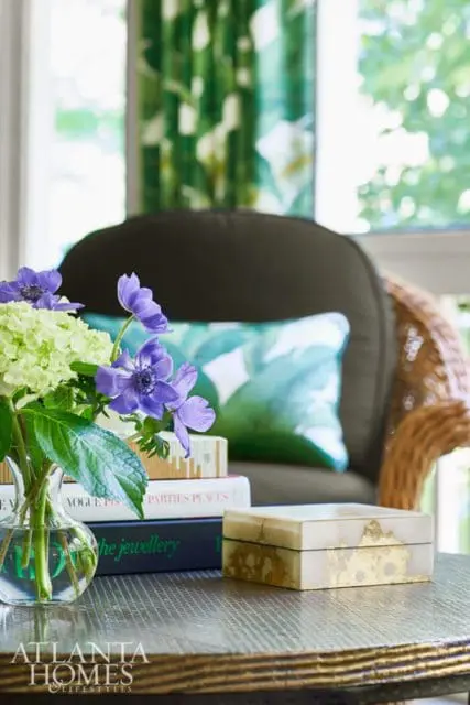 Dark upholstered chair with wicker arms, tropical print pillow, and a glass vase of purple and green flowers on a table with books and a decorative box.