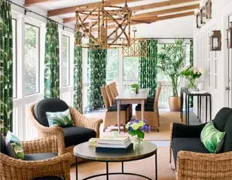Sunroom with wicker chairs and sofas upholstered in black fabric, tropical leaf print curtains, and bamboo pendant lights over a dining table.