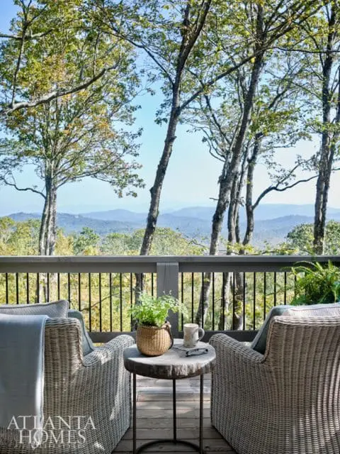 Wicker armchairs on a wooden deck with a view of mountains and trees. A small table holds a potted plant, mug, and glasses.