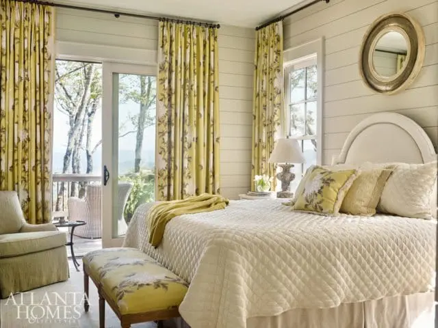 Bedroom with a queen-sized bed, white quilted comforter, yellow floral curtains, and a green armchair. A sliding glass door opens to a balcony with a view of trees and mountains.