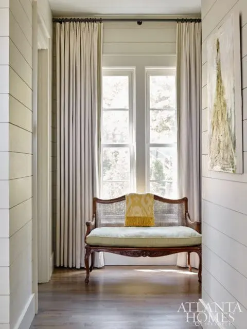 Hallway with shiplap walls, dark wood floors, and a vintage cane-backed bench with a light green cushion and yellow throw. Tall curtains frame a double window.