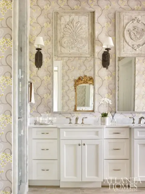 White bathroom vanity with marble countertops, brass faucets, and floral wallpaper. Ornate carved wood panels hang above the mirrors.