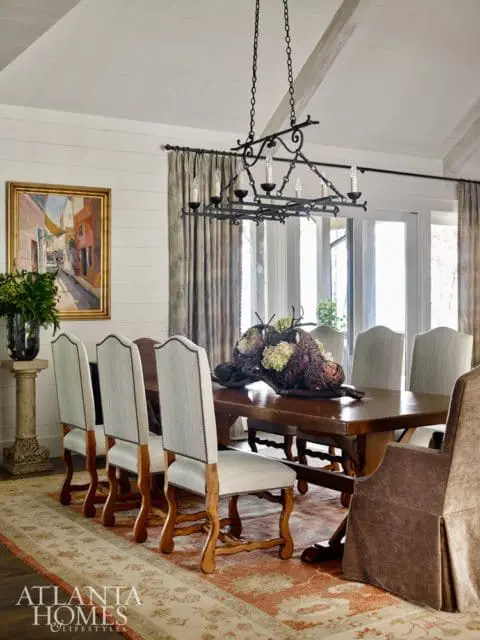 Dining room with a wooden table and chairs, a large ornate chandelier, and a painting on the wall. The room features a vaulted ceiling and large windows.