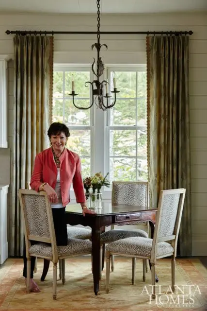 Woman in coral jacket standing by a dark wood dining table with leopard print upholstered chairs, beneath a wrought iron chandelier.
