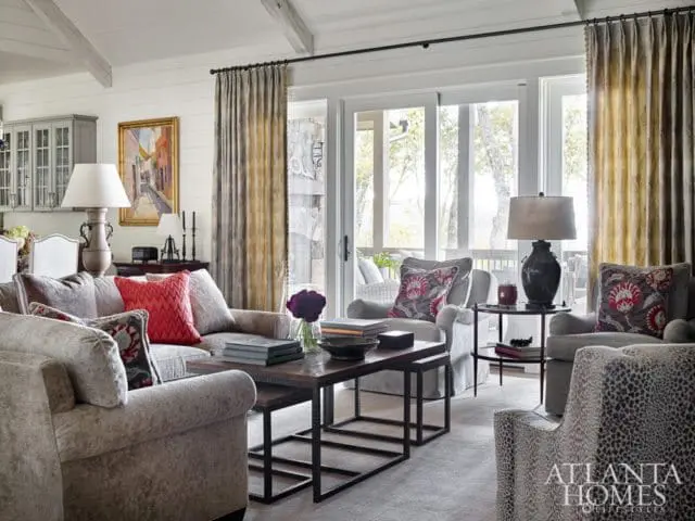 Living room with plush gray sofas and armchairs, patterned accent pillows, dark wood coffee table, and large windows overlooking a wooded area.