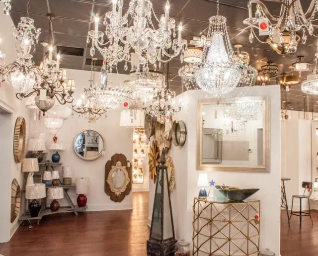 Showroom interior with numerous crystal chandeliers hanging from a dark ceiling, and a display of lamps and decorative mirrors on the walls and floor.
