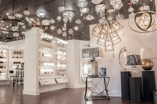 Showroom interior with a wide selection of ceiling and table lamps, including a large geometric chandelier and a mirrored table lamp with a silver shade.