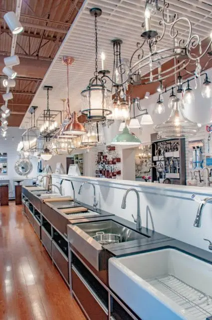 Showroom display of various kitchen sinks and faucets, with a selection of pendant lights and chandeliers hanging overhead.