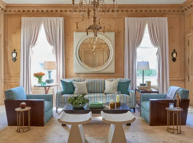 Living room with wood-paneled walls, featuring a striped sofa, two blue patterned armchairs, a dark wood coffee table with white stools, and a large round mirror above the sofa.