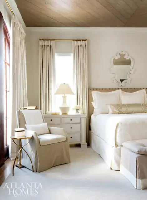Bedroom with a white upholstered armchair, a white dresser with gold hardware, and a bed with white linens. The ceiling features wood planks.