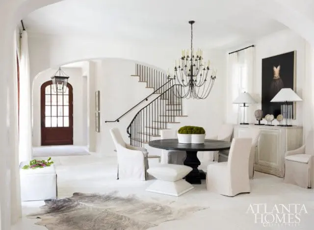 White entryway and dining area with a dark wood arched door, curved staircase with black railing, round dark wood dining table with white slipcovered chairs, and a large black framed artwork above a distressed white sideboard.