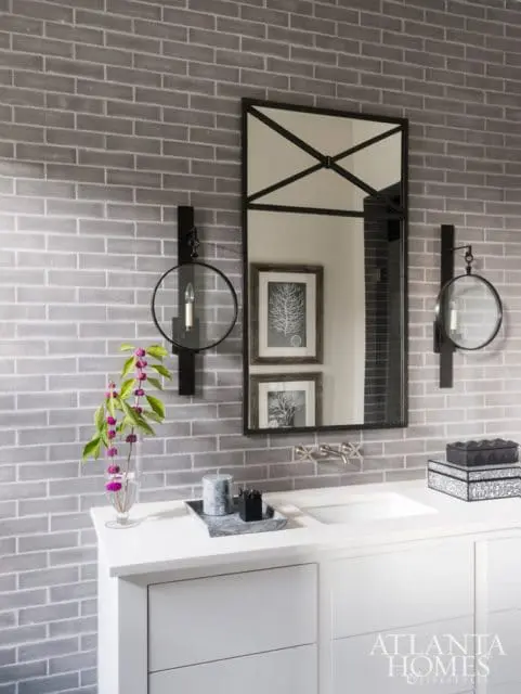 Modern bathroom vanity with white countertop and white drawers, featuring a white sink, brushed nickel faucet, and gray brick tile backsplash. A tall black framed mirror hangs above the sink, flanked by two circular sconces.