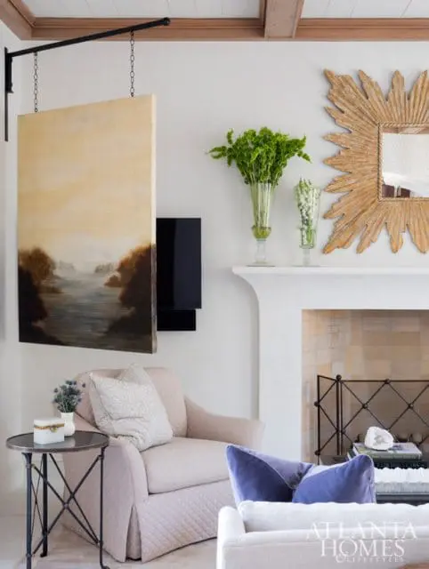 Living room with a quilted armchair, a landscape painting hanging from a metal arm, a sunburst mirror over a fireplace, and a coffee table with books and a geode.