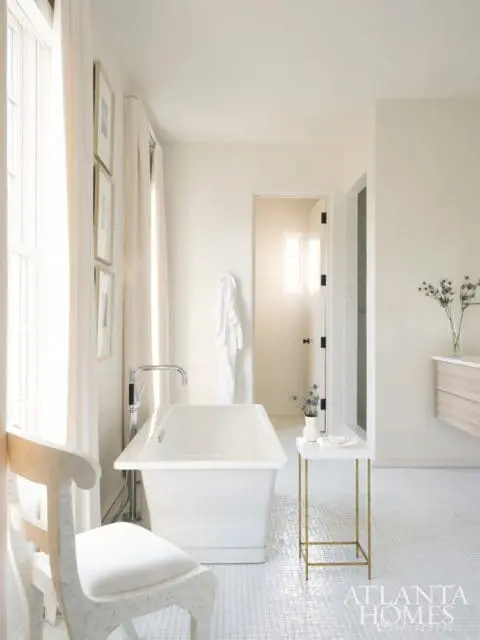 Creamy white bathroom with a freestanding soaking tub, white mosaic tile floor, a decorative chair, and a small gold side table with a vase of flowers.