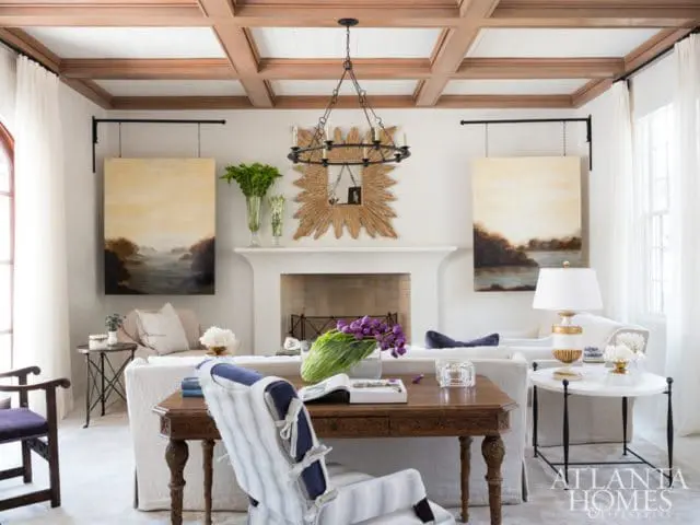 Living room with coffered wood ceiling, white fireplace, and two landscape paintings flanking a sunburst mirror.