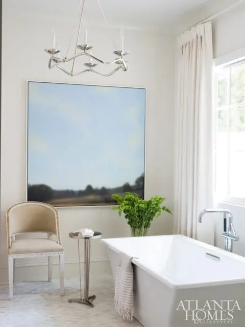 Elegant bathroom with a freestanding white tub, a plush upholstered chair, abstract landscape art, and a modern chandelier.