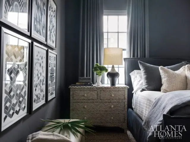 Dark gray bedroom with a black upholstered bed, patterned nightstand, framed abstract art, and gray curtains.