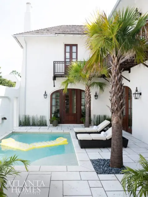 White stucco house with dark wood doors and balconies, featuring a small pool with a yellow float, two lounge chairs, and palm trees.