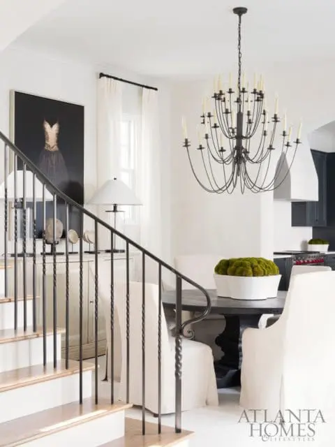 Dining area with a dark wood table, white slipcovered chairs, and a large black chandelier. A staircase with a metal railing is in the foreground.