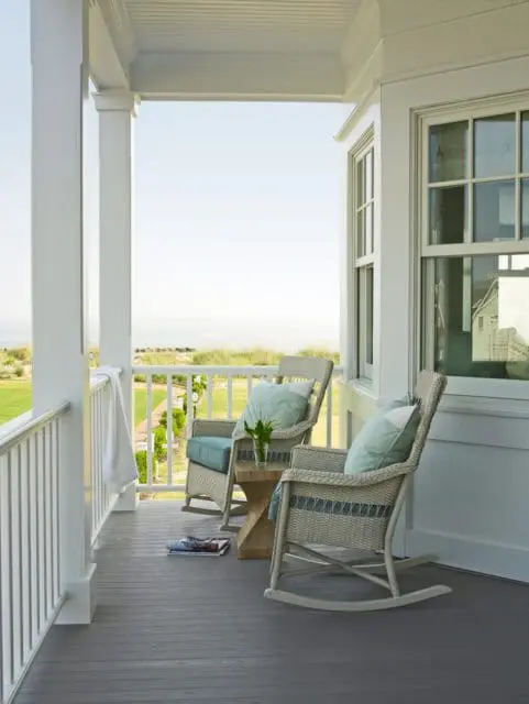 White porch with gray floorboards, two wicker rocking chairs with blue cushions, and a view of the ocean.