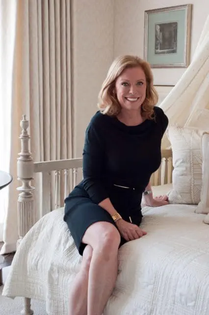 Woman with blonde hair wearing a black dress and gold bracelet sits on a white quilted bed with decorative pillows.