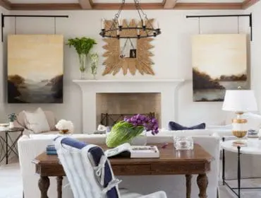 Living room with white fireplace, two landscape paintings, a sunburst mirror, and a wooden desk with a striped chair.