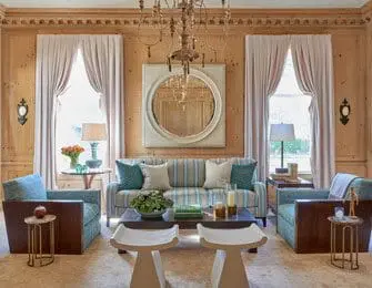 Living room with wood-paneled walls, a striped sofa, two blue armchairs, and a large round mirror above the sofa.
