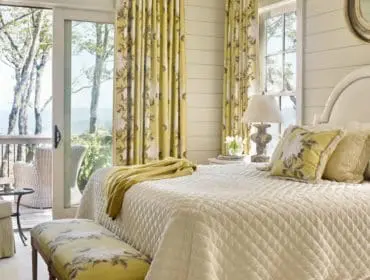 Bedroom with a white quilted bed, yellow floral curtains, and a green armchair. A sliding glass door opens to a balcony with a view of trees and water.