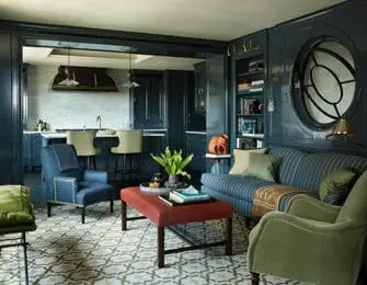 Living room with navy blue walls and built-in shelving, featuring a striped sofa, two armchairs, a red ottoman, and a patterned rug. An open doorway reveals a kitchen with a marble backsplash and island seating.