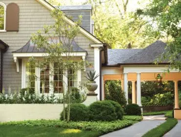 Shingle-sided house with arched window and decorative shutter, flanked by manicured hedges and a covered walkway with columns.