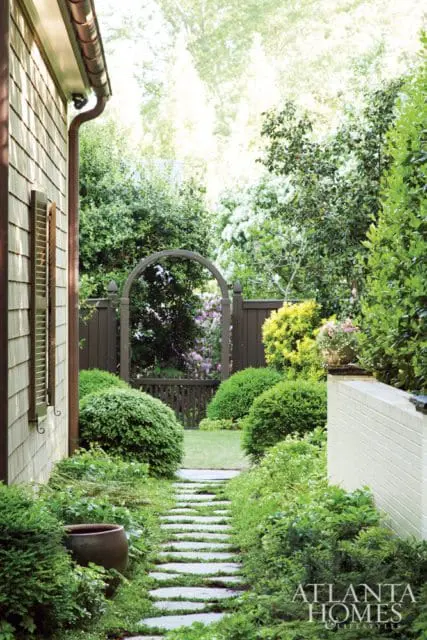 Stone pathway through a lush garden with manicured bushes, a wooden arbor, and a white brick wall. The Atlanta Homes & Lifestyles logo is visible in the lower right corner.