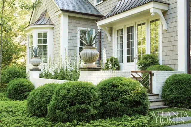 Shingle-sided house with bay window and double doors, flanked by manicured bushes and white brick planters with agave plants.