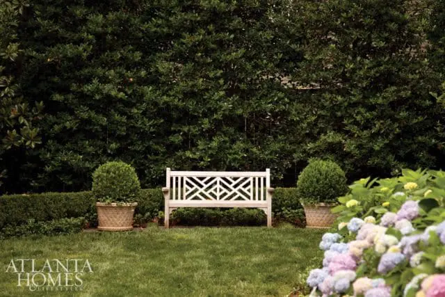 A white wooden bench with a geometric pattern on its back sits in a garden with manicured hedges, potted topiaries, and blooming hydrangeas.