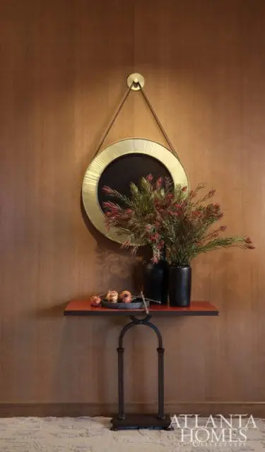 Wood paneled wall with a round gold-trimmed mirror hanging above a console table holding black vases with red-tipped greenery and a bowl of pomegranates.