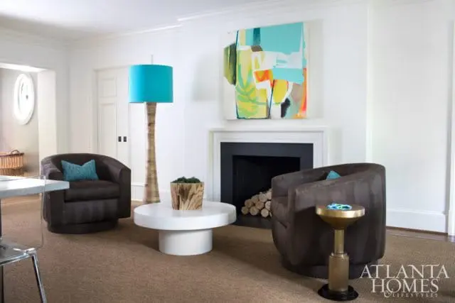 Living room with two dark brown swivel chairs, a white pedestal coffee table, a tall floor lamp with a teal shade, and a modern abstract painting above a black fireplace.
