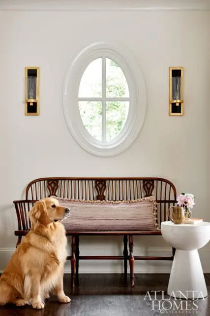 Entryway with a golden retriever sitting next to a dark wood spindle bench topped with a striped pillow, beneath an oval window and flanked by gold sconces.