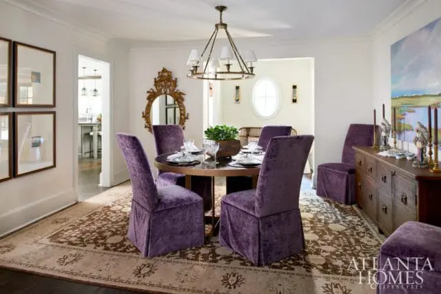 Dining room with plush purple velvet chairs around a round table, ornate gold mirror, and a landscape painting above a wooden buffet.
