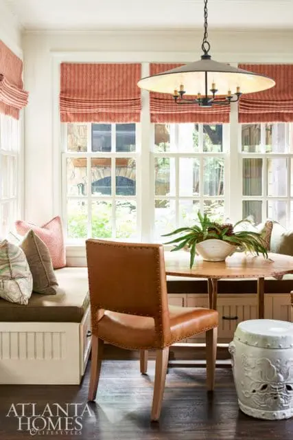 Breakfast nook with built-in banquette seating, a round wood table, leather chair, and patterned Roman shades.