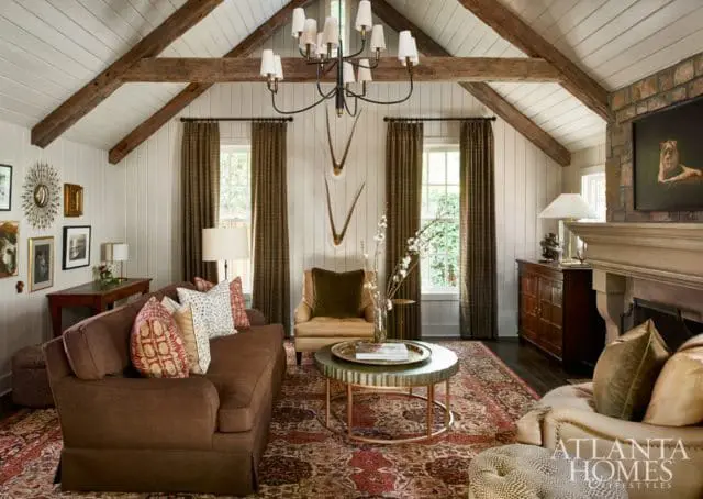 Living room with vaulted ceiling, exposed wooden beams, stone fireplace, brown sofa, patterned rug, and decorative wall art.