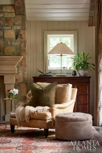 Living room with leather armchair, stone fireplace, dark wood cabinet, and patterned ottoman. A window overlooks greenery.