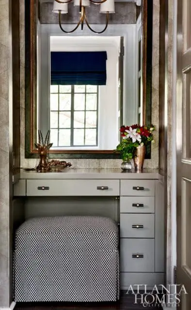 Gray vanity with black and white patterned ottoman, brass-framed mirror reflecting a window with a blue Roman shade, and a vase of flowers.